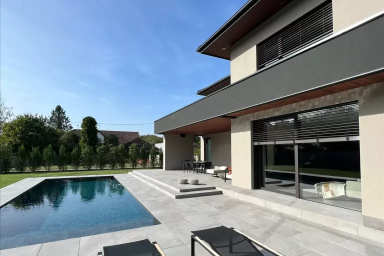 Façade d’une maison contemporaine avec grandes baies vitrées, brise-soleil à lames, terrasse partiellement couverte et piscine.