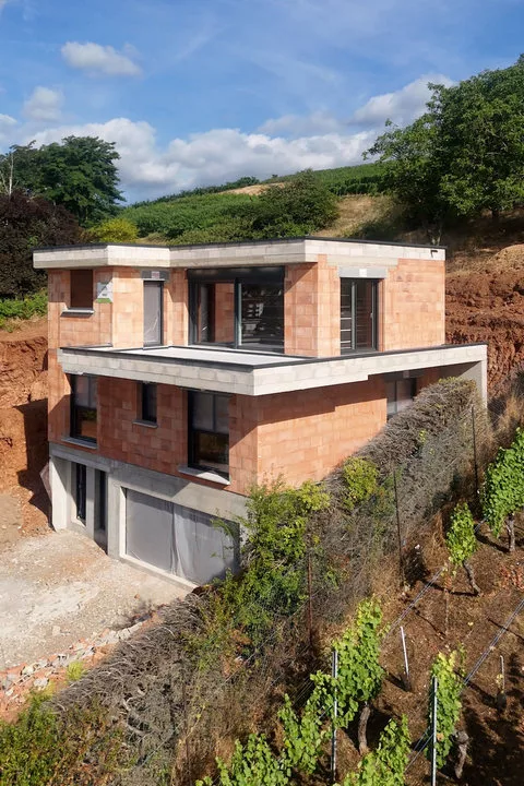 Maison en brique en cours de construction sur un terrain en pente, avec toitures plates et garage au niveau inférieur.
