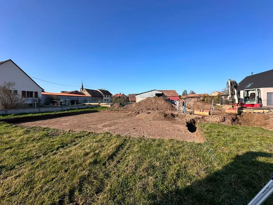 Terrain en cours de terrassement pour une maison individuelle, avec une pelle mécanique et des repères d’implantation.