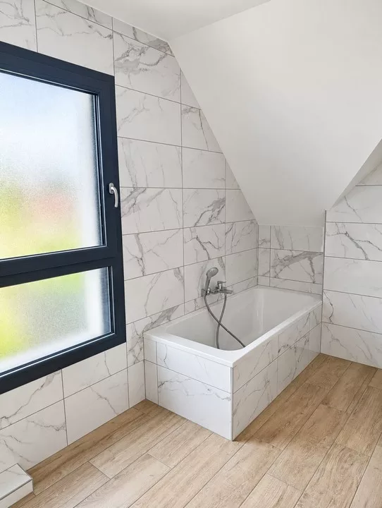 Salle de bains sous pente avec baignoire, carrelage effet marbre, sol imitation bois et fenêtre anthracite.