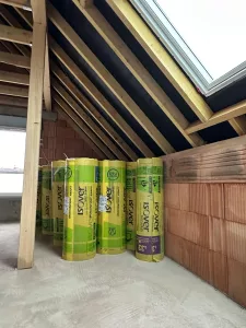 Rouleaux d’isolant Isover stockés sous une charpente en bois, près d’un mur en briques et d’une fenêtre de toit.